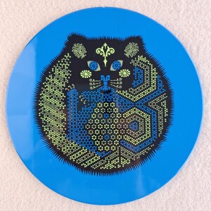 image shows a set of 6 Reginald Corfield black cat placemats, each is 16.5cm diameter circular metal & cork coasters with striking psychedelic design. The photos are taken against a stone and pink background.