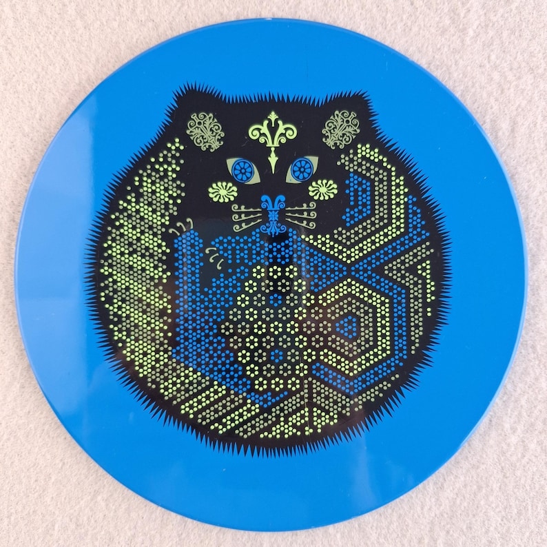 image shows a set of 6 Reginald Corfield black cat placemats, each is 16.5cm diameter circular metal & cork coasters with striking psychedelic design. The photos are taken against a stone and pink background.