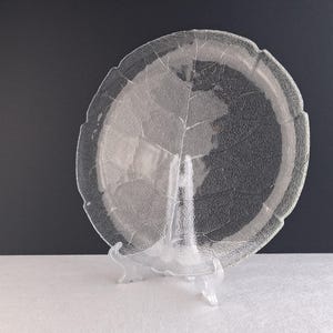 May include: A clear glass plate with a leaf design, on a clear plastic stand. The plate has a textured surface and a scalloped edge. The background is dark grey.