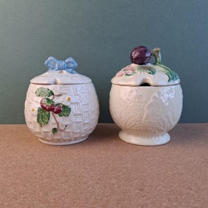 May include: Two cream-coloured ceramic jars with lids. One has a woven basket design, a blue bow on the lid, and a berry branch. The other has a textured surface with a purple fruit on the lid. Both jars are cream-coloured.