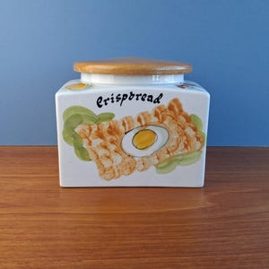 May include: A white ceramic crispbread jar with a wooden lid. The jar has a hand-painted illustration of crispbread and an egg, with the word "Crispbread" in black script. The jar is rectangular and sits on a wooden surface.