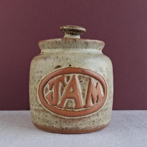 May include: A ceramic jam jar with a lid, featuring a speckled beige and brown glaze. The word "JAM" is embossed in a reddish-brown oval on the front. The jar has a rounded shape and a small handle on the lid.