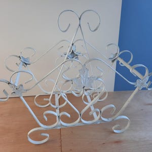 May include: White metal magazine rack with a decorative scroll design. The rack has a triangular shape and features leaf-shaped accents.