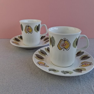 May include: Two white ceramic coffee cups and saucers with a mid-century modern design. Each cup and saucer features a leaf and fruit pattern in yellow, brown, and green. The cups have a handle and are set on a grey surface against a pink background.