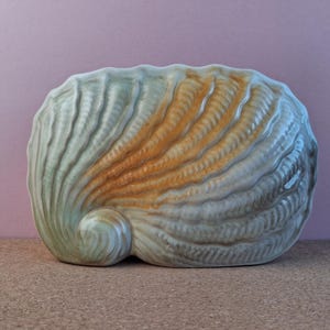 May include: A ceramic seashell sculpture in pale green and orange. The sculpture has a textured surface with detailed ridges and a rounded, spiral shape. The background is a solid purple colour.