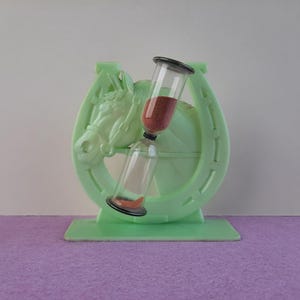 May include: A green plastic hourglass shaped like a horseshoe with a horse's head on the side. The hourglass is filled with red sand.