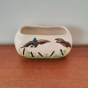 May include: A white ceramic planter with a rectangular shape and a raised design of two ducks flying over a bed of green reeds.