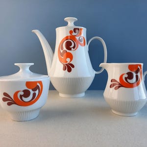 May include: A white ceramic tea set with an orange and brown swirl design. The set includes a teapot, creamer, and sugar bowl.