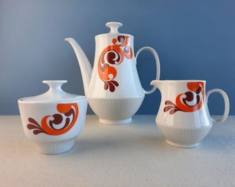 May include: A white ceramic tea set with an orange and brown swirl design. The set includes a teapot, creamer, and sugar bowl.