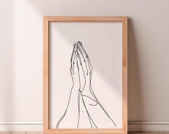 Op de afbeelding: Een minimalistische kunstprint in een lijst met een doorlopende lijntekening van twee handen die in gebed gevouwen zijn. Het kunstwerk is in zwart op een crèmekleurige achtergrond geplaatst, in een natuurlijke houten lijst.