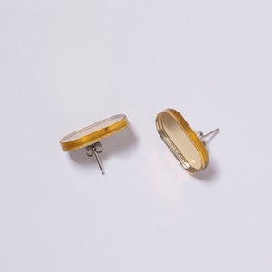 Może przedstawiać: Para owalnych kolczyków z pozłacanym metalowym obramowaniem i białym środkiem. Kolczyki mają zapięcie na sztyft.