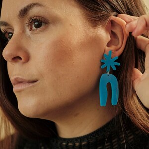 May include: Turquoise blue abstract earrings. The earrings feature a starburst shape at the top and a curved, U-shaped design below. The earrings are worn with a black mesh top.