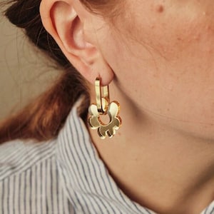 Puede incluir: Primer plano de una mujer que lleva un pendiente dorado con forma de flor. El pendiente es de estilo aro con un diseño floral. La mujer lleva una camisa abotonada a rayas.