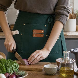 May include: A dark green apron with two brown leather accents and brass rivets. The apron has a pocket on the front and is tied at the waist. A person is shown cutting a red onion on a wooden cutting board.