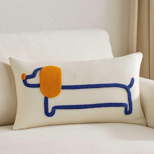 May include: Cream-colored rectangular decorative pillow featuring a blue outline of a dachshund dog with an orange ear. The pillow is placed on a white sofa, creating a cozy and inviting aesthetic.