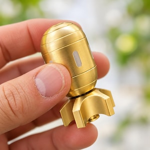 Op de afbeelding: Een cilindervormige messing fidget spinner met een geborstelde metalen afwerking, voorzien van horizontale groeven langs het afgeronde bovendeel en een stervormige basis met zes duidelijke vinnen. Kleine, rechthoekige witte inzetstukken zijn in de behuizing verwerkt.