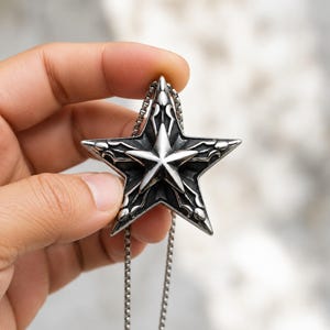 Puede incluir: Un colgante de estrella de metal plateado con un diseño en capas que presenta una estrella más pequeña y pulida centrada sobre una estrella más grande y oscura con detalles intrincados y elevados. El colgante cuelga de una cadena fina.