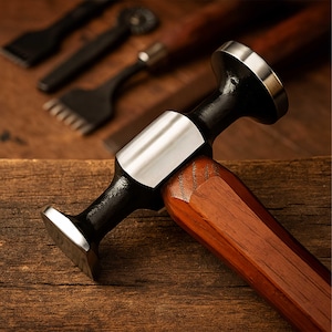 Puede incluir: Primer plano de un martillo de marroquinería con mango de madera y cabeza negra con superficie de golpeo plateada. Otras herramientas de marroquinería están en el fondo. El martillo está sobre una superficie de madera.