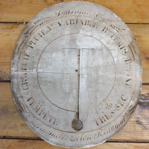 May include: A white, oval-shaped weather instrument with a black needle and markings. The instrument is labeled "GRALCHE PLUIE VARIABLE TEMPS TRES-SEC" and "Chermometre Sclon Reaumur".