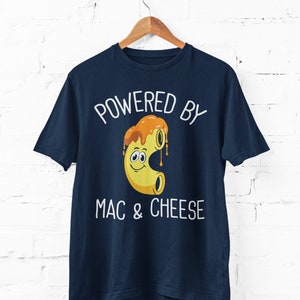 May include: Navy blue t-shirt with the text "POWERED BY MAC & CHEESE" in white. The graphic features a smiling cartoon macaroni noodle covered in cheese sauce. The shirt is hanging on a wooden hanger.