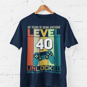Op de afbeelding: Een marineblauw T-shirt met een retro-grafisch ontwerp. De grafiek toont een videogamecontroller, de tekst "Level 40" en de woorden "40 Years of Being Awesome Unlocked Press Start to Continue".