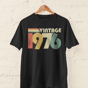 May include: A black short-sleeve t-shirt featuring a graphic print with the text VINTAGE 1976. The design includes a distressed, multi-colored horizontal stripe pattern above the year, utilizing muted shades of red, yellow, green, and blue.