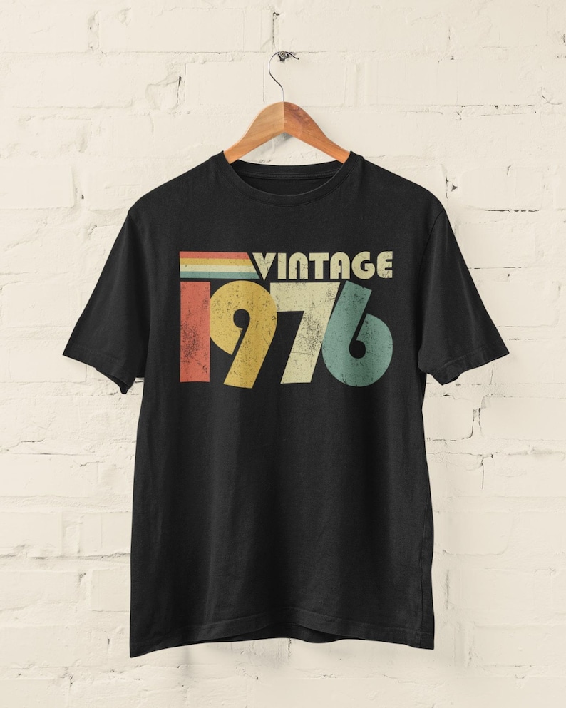 May include: A black short-sleeve t-shirt featuring a graphic print with the text VINTAGE 1976. The design includes a distressed, multi-colored horizontal stripe pattern above the year, utilizing muted shades of red, yellow, green, and blue.