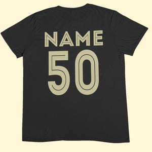 May include: A black short-sleeved t-shirt featuring the word NAME printed in large beige capital letters above the number 50 in a matching beige font. The design is centered on the back of the garment, utilizing a clean, bold, sans-serif typeface.