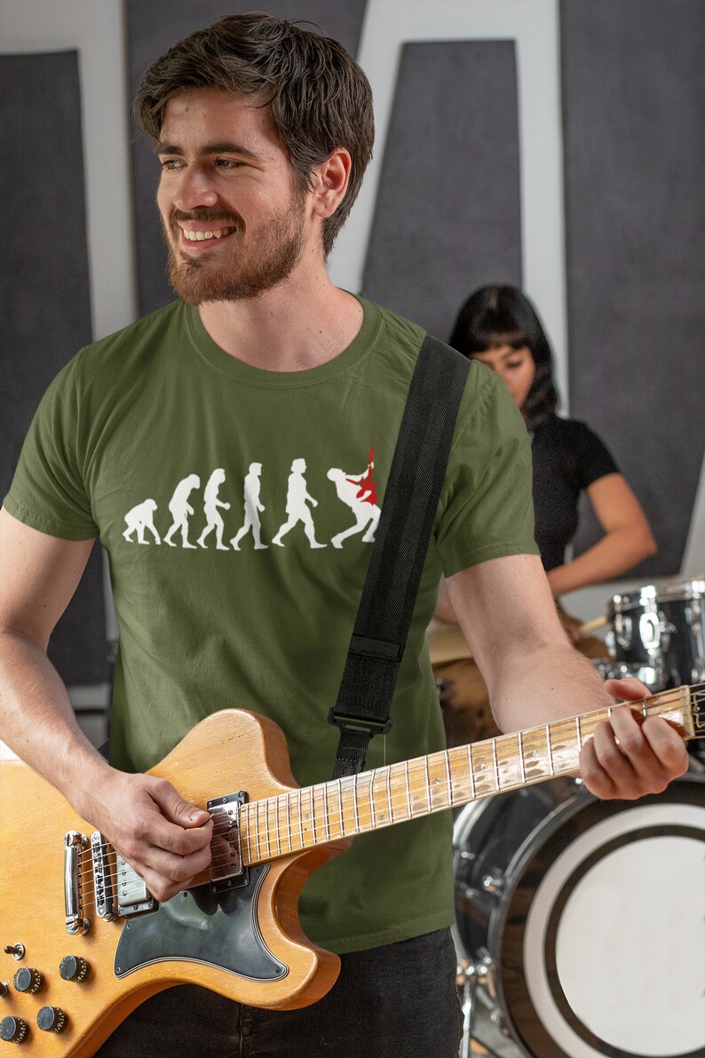 May include: A man wearing a green t-shirt with a graphic of the evolution of man culminating in a person playing an electric guitar. He is holding a brown electric guitar and smiling.