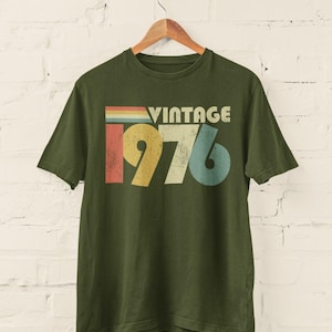 May include: An olive green short-sleeve t-shirt featuring a graphic print with the text VINTAGE 1976. The design includes horizontal stripes in red, yellow, and blue above the year, with the numbers 1976 displayed in a distressed, multi-colored font.