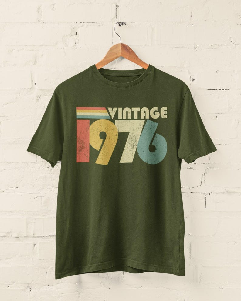 May include: An olive green short-sleeve t-shirt featuring a graphic print with the text VINTAGE 1976. The design includes horizontal stripes in red, yellow, and blue above the year, with the numbers 1976 displayed in a distressed, multi-colored font.
