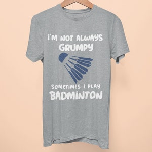 Op de afbeelding: Een grijs T-shirt met de tekst "Ik ben niet altijd chagrijnig, soms speel ik badminton" en een blauwe badminton shuttlecock afbeelding.