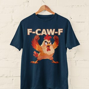 May include: Navy blue t-shirt featuring a cartoon chicken with raised arms and an angry expression. The text "F-CAW-F" is printed above the chicken in a bold, retro font. The shirt is hanging on a wooden hanger against a white brick wall.