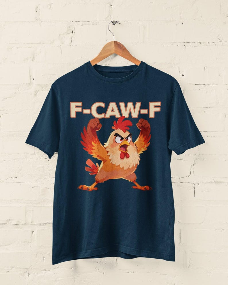 May include: Navy blue t-shirt featuring a cartoon chicken with raised arms and an angry expression. The text "F-CAW-F" is printed above the chicken in a bold, retro font. The shirt is hanging on a wooden hanger against a white brick wall.