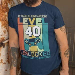 Op de afbeelding: Een marineblauw T-shirt met een retro-grafische afbeelding waarop staat "40 Years of Being Awesome Level 40 Unlocked Press Start to Continue". De afbeelding toont een videogamecontroller en een kleurrijke achtergrond.