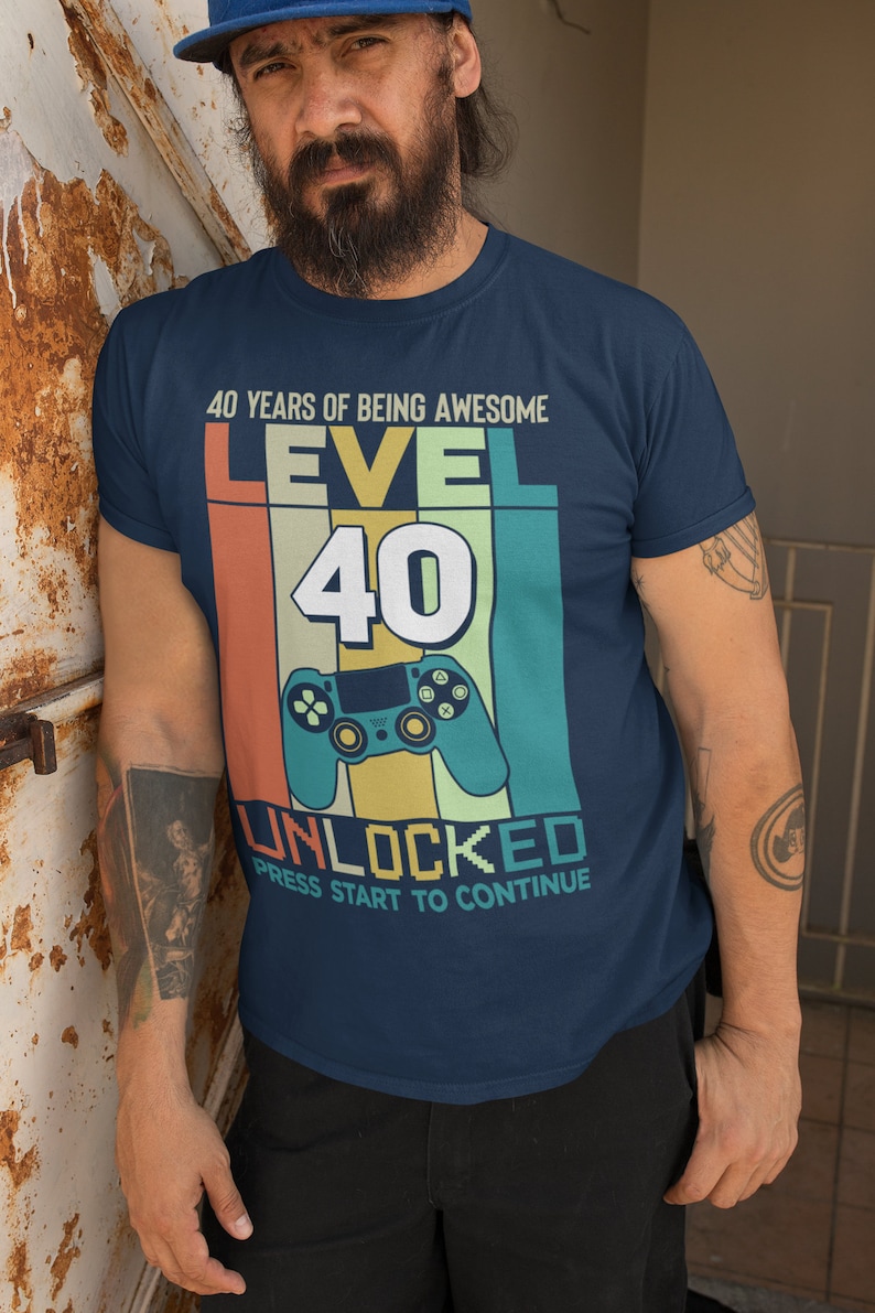 Op de afbeelding: Een marineblauw T-shirt met een retro-grafische afbeelding waarop staat "40 Years of Being Awesome Level 40 Unlocked Press Start to Continue". De afbeelding toont een videogamecontroller en een kleurrijke achtergrond.