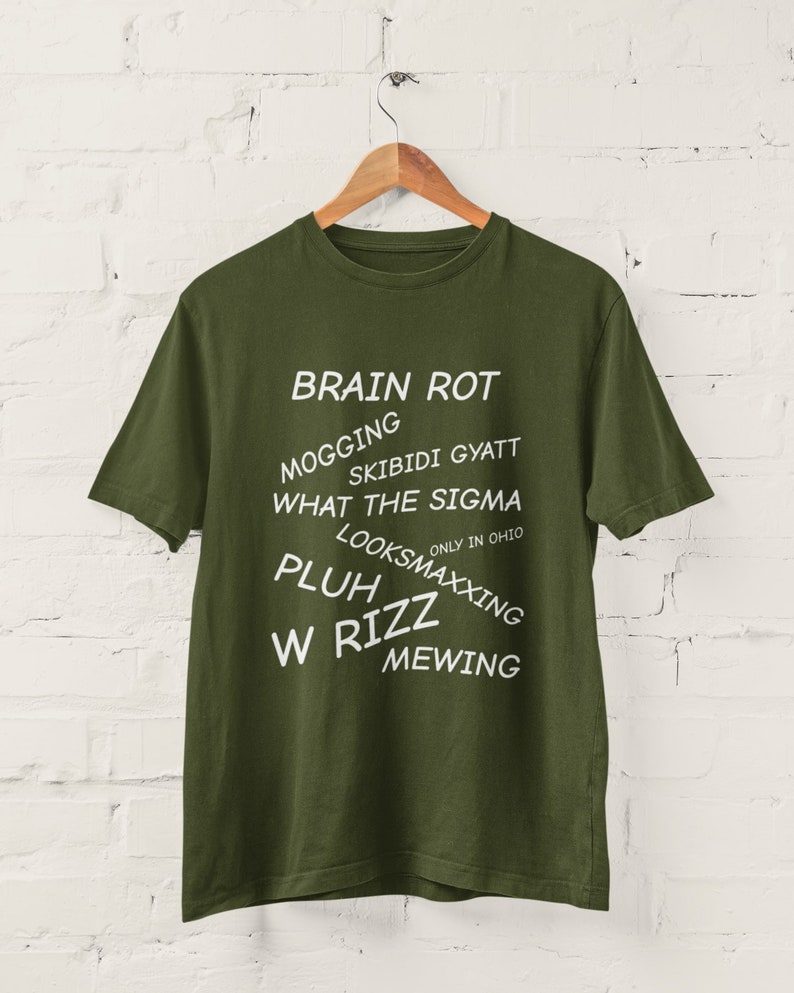 May include: A green t-shirt with white text that reads "BRAIN ROT", "MOGGING", "SKIBIDI GYATT", "WHAT THE SIGMA", "ONLY IN OHIO", "LOOKSMAXXING", "PLUH", "W RIZZ", and "MEWING".