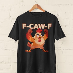 May include: Black t-shirt featuring a cartoon chicken with raised arms and an angry expression. The text "F-CAW-F" is printed above the chicken in a bold, orange font. The shirt is hanging on a wooden hanger.