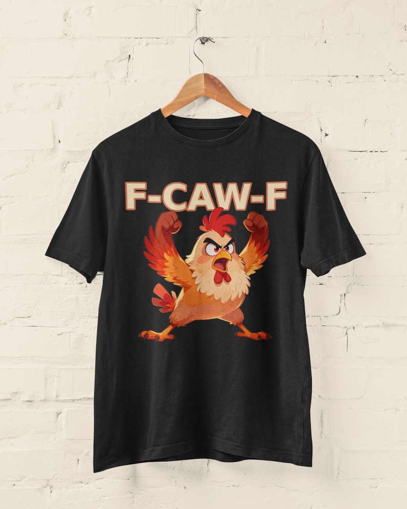May include: Black t-shirt featuring a cartoon chicken with raised arms and an angry expression. The text "F-CAW-F" is printed above the chicken in a bold, orange font. The shirt is hanging on a wooden hanger.