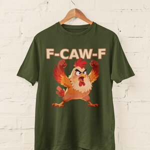 May include: Olive green t-shirt featuring a cartoon chicken with raised wings and an angry expression. The text "F-CAW-F" is printed above the chicken in a bold, orange font. The shirt is hanging on a wooden hanger.