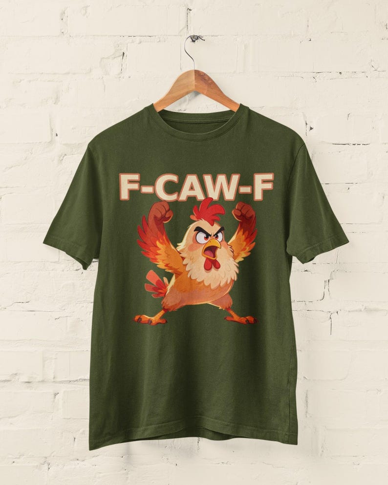 May include: Olive green t-shirt featuring a cartoon chicken with raised wings and an angry expression. The text "F-CAW-F" is printed above the chicken in a bold, orange font. The shirt is hanging on a wooden hanger.