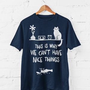 May include: Navy blue t-shirt featuring a white graphic of a cat knocking over a vase, a picture frame and an alarm clock. The text "This is why we can't have nice things" is printed below the graphic.