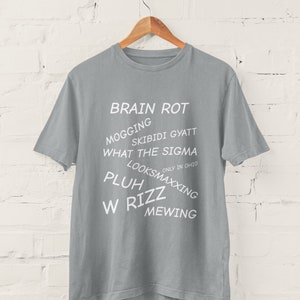 May include: A gray t-shirt with white text that reads "BRAIN ROT", "MOGGING", "SKIBIDI GYATT", "WHAT THE SIGMA", "ONLY IN OHIO", "LOOKSMAXXING", "PLUHZZ", "W RIZZ", and "MEWING".
