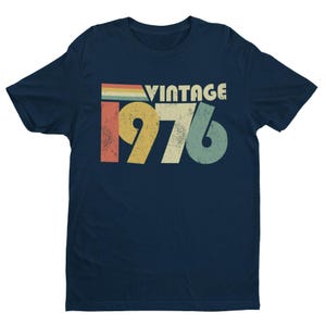 May include: A navy blue short-sleeve t-shirt featuring a distressed graphic print on the chest. The design includes the word VINTAGE above the year 1976 in large, bold, multi-colored numbers with a horizontal striped bar in red, yellow, and blue.