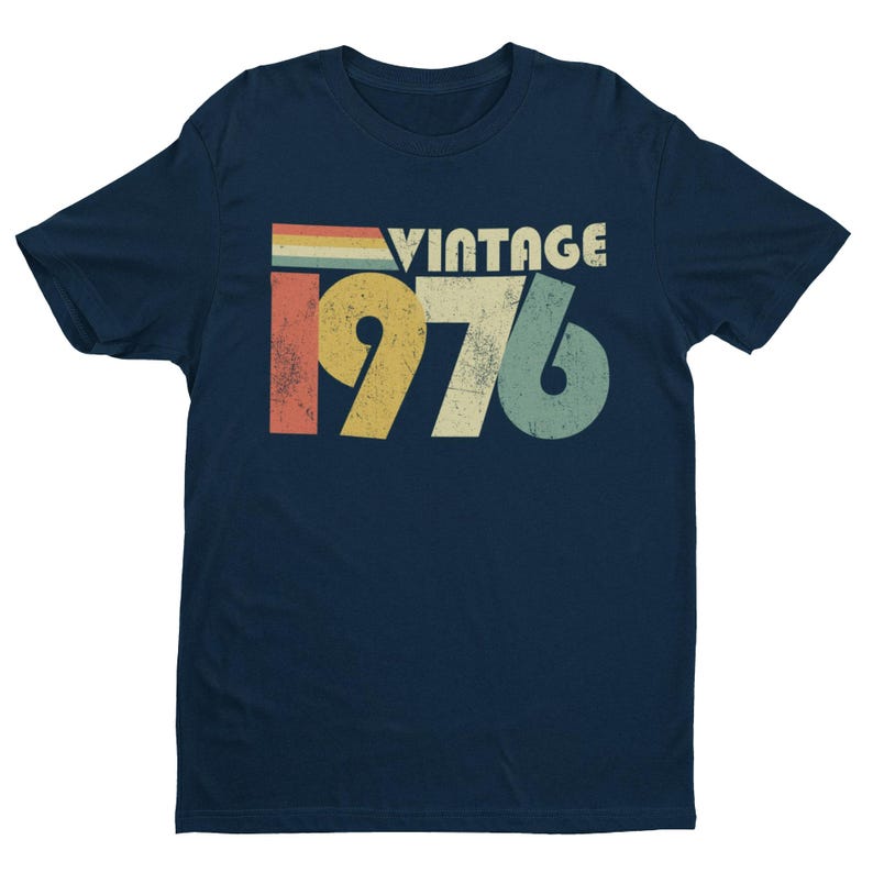 May include: A navy blue short-sleeve t-shirt featuring a distressed graphic print on the chest. The design includes the word VINTAGE above the year 1976 in large, bold, multi-colored numbers with a horizontal striped bar in red, yellow, and blue.