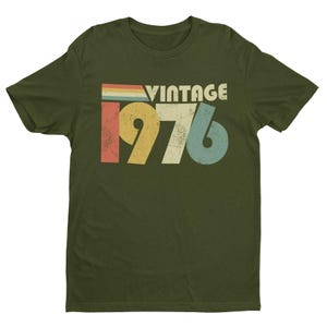 May include: An olive green short-sleeve t-shirt featuring a distressed graphic print on the chest. The design includes the word VINTAGE in white capital letters above the large numbers 1976, rendered in yellow, beige, and blue with a retro striped accent.