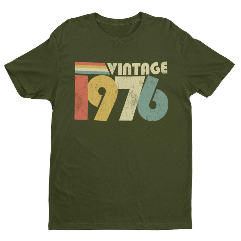 May include: An olive green short-sleeve t-shirt featuring a distressed graphic print on the chest. The design includes the word VINTAGE in white capital letters above the large numbers 1976, rendered in yellow, beige, and blue with a retro striped accent.