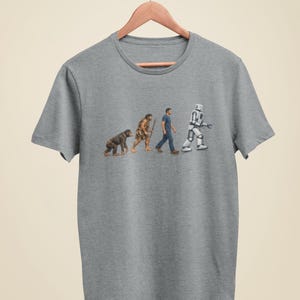 Peut inclure: T-shirt gris avec un motif humoristique sur l'évolution. Le design représente une progression du singe à l'homme des cavernes, puis à l'homme moderne, et enfin à un Stormtrooper. Le t-shirt est suspendu à un cintre en bois.