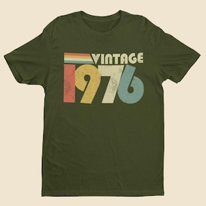 May include: An olive green short-sleeve t-shirt featuring a distressed graphic print on the chest. The design includes the word VINTAGE in white letters above the large numbers 1976, which are colored in yellow, off-white, and light blue.