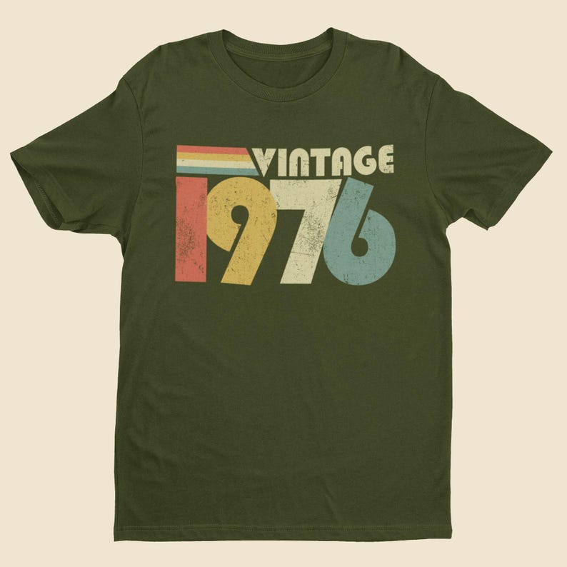 May include: An olive green short-sleeve t-shirt featuring a distressed graphic print on the chest. The design includes the word VINTAGE in white letters above the large numbers 1976, which are colored in yellow, off-white, and light blue.